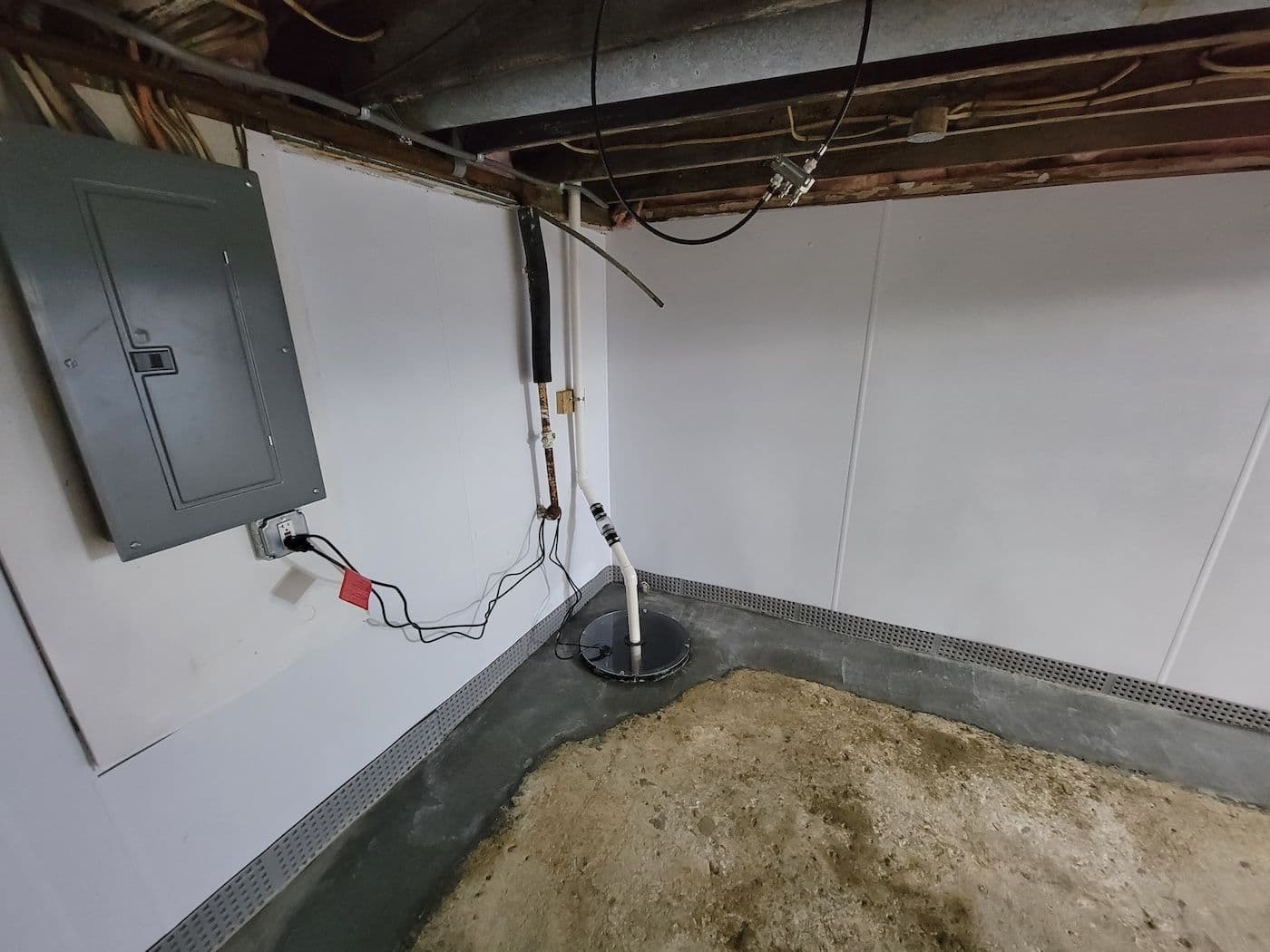 Basement sump pump pit with submersible pump, discharge pipes, electrical panel on wall, and metal drain basin surrounded by concrete floor.