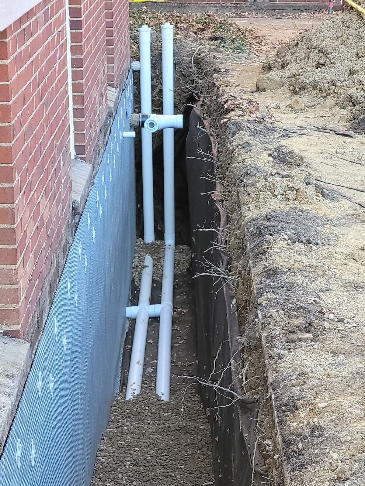 PVC pipes and black conduit installed along foundation of brick building next to excavated trench with exposed soil and roots.